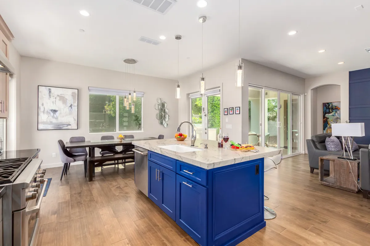 An open-plan kitchen and dining area featuring a kitchen island with blue cabinetry, a nearby dining table, large windows letting in natural light, and contemporary light fixtures. The seamless flow between kitchen and dining spaces invites family interaction and group meals.