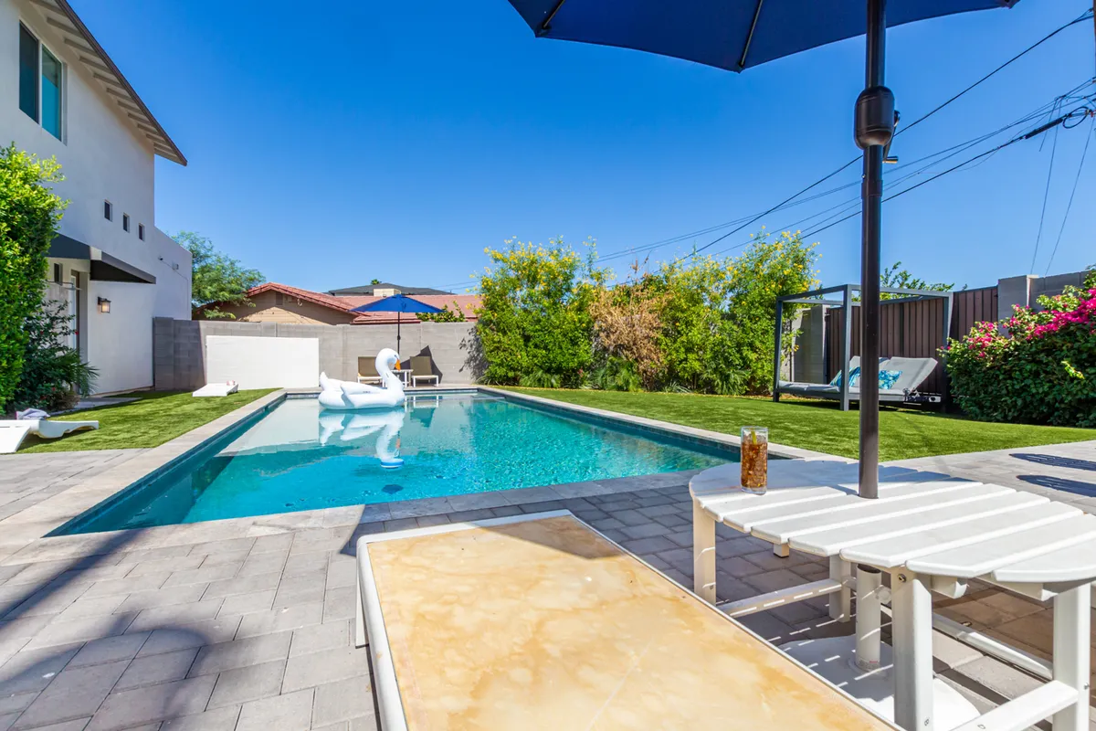 A vibrant backyard scene featuring a sparkling swimming pool bordered by lounge chairs, umbrellas, lush greenery, and a decorative inflatable swan. The area looks welcoming for both relaxation and group activities.