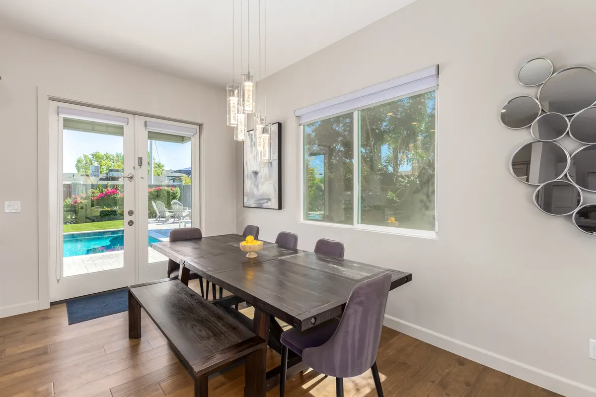 A stylish dining room with a large window overlooking the pool, a sophisticated chandelier, and a decorative mirror. The central dining table is ready for gatherings, with an inviting atmosphere for both sunlit breakfasts and intimate dinners.