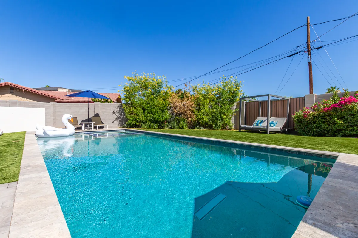 A vibrant backyard scene featuring a sparkling swimming pool with an inflatable swan, surrounded by plenty of lounge chairs and lush greenery. The area exudes a playful, social atmosphere ideal for gathering with friends to relax, sunbathe, or host a pool party.