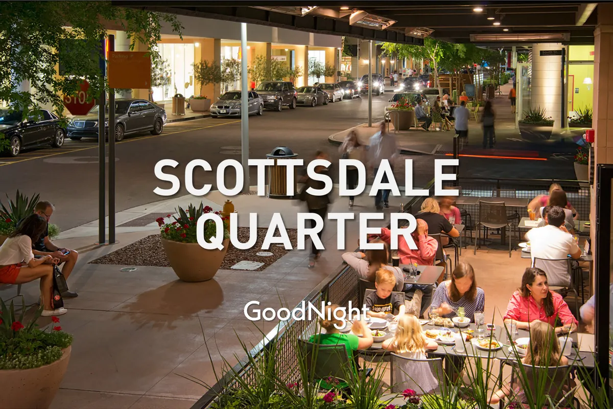 A lively outdoor dining area with people socializing under glowing street lights. The scene captures the vibrant atmosphere of an urban neighborhood, where guests are enjoying food, drinks, and conversation in the open air.