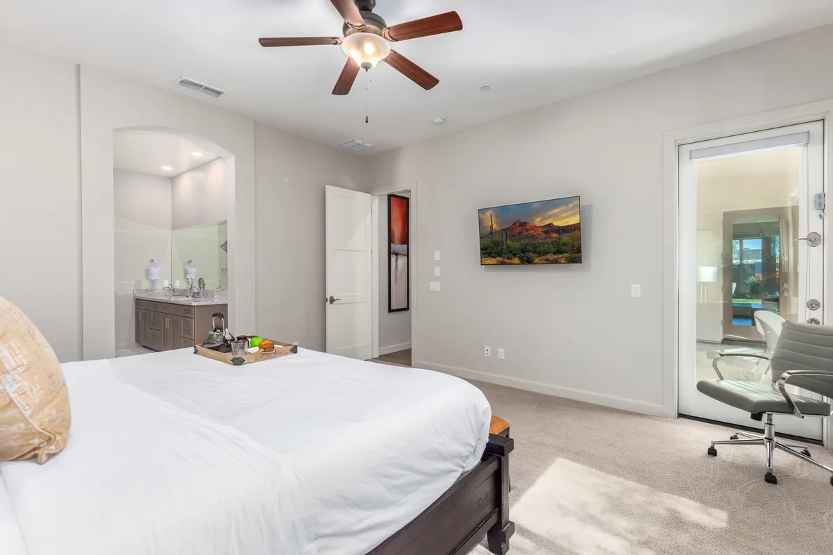 A tranquil bedroom with a king-sized bed topped with a decorative tray, wall-mounted TV, and a door opening to an outdoor area. Natural light fills the room, creating a cozy and restful atmosphere.