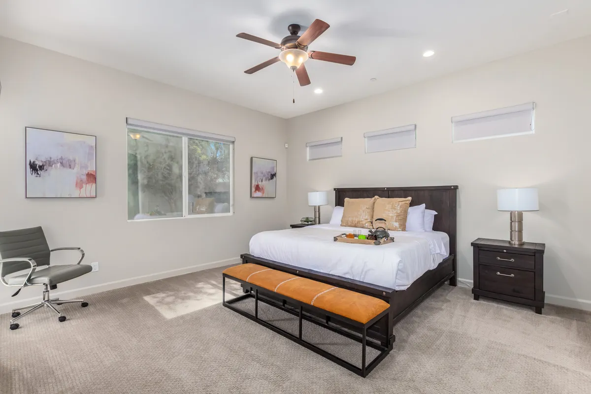 A bright and tranquil bedroom with a plush bed, decorative pillows and throw, a window letting in natural light, stylish wall art, and a ceiling fan. The space feels peaceful and inviting.