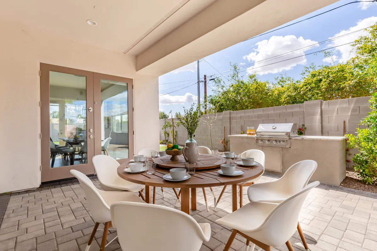 An inviting outdoor dining area with a large table, modern grill, and sliding glass doors opening to the greenery beyond. The setup is ideal for al fresco meals, group breakfasts, or evening socials, connecting indoor and outdoor living spaces.