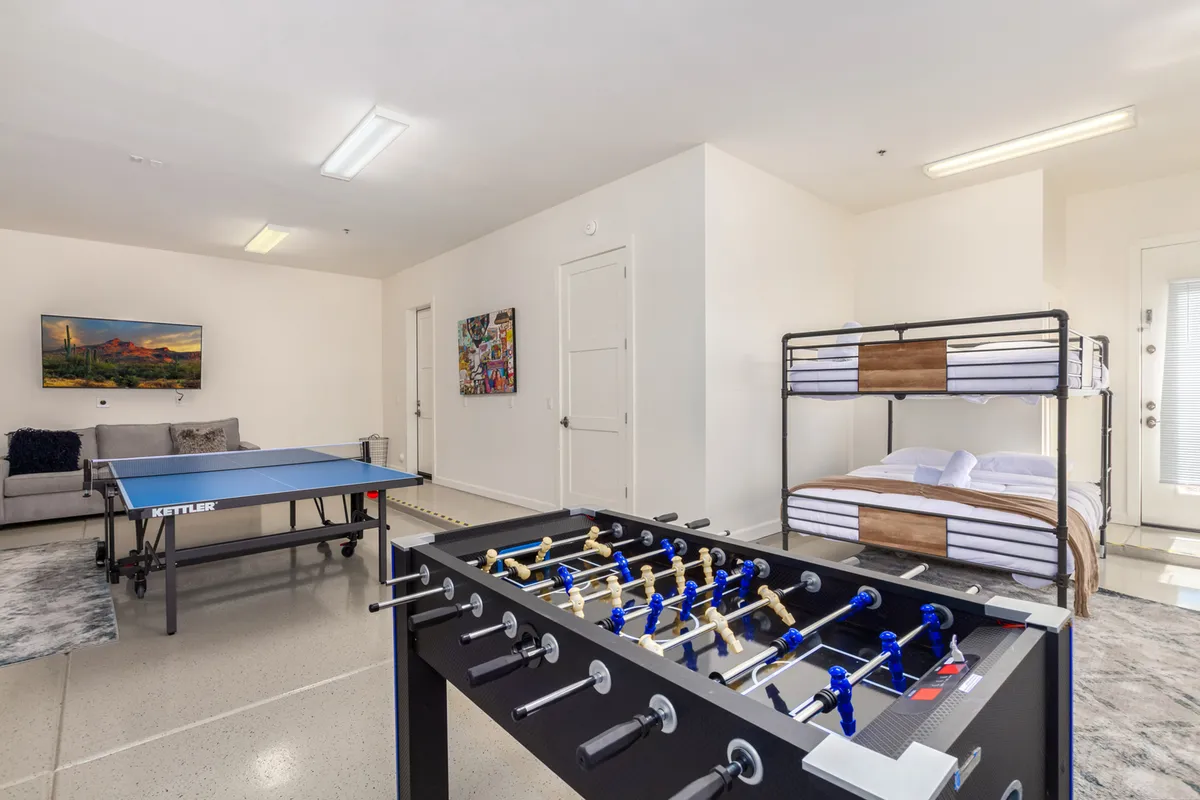 A vibrant bunk/game room featuring four bunk beds, a foosball table, a ping pong table, a comfy sofa, and colorful wall art. The space is designed for both sleep and play, with a youthful, energetic vibe.