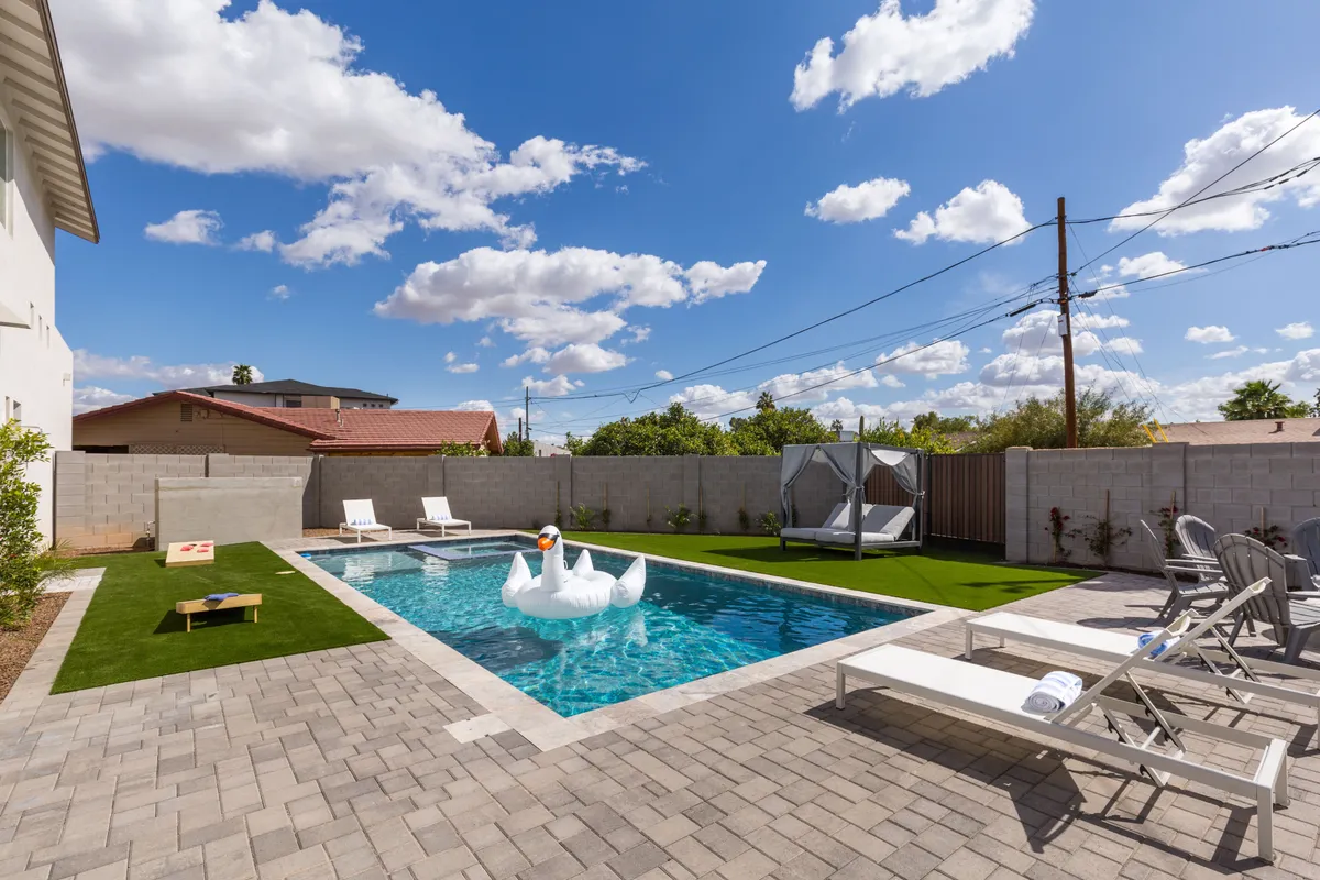 This exterior shot highlights a vibrant backyard scene featuring a large swimming pool, an inflatable swan, multiple lounge chairs, a hammock, and a visible cornhole game setup. The area is surrounded by lush greenery, creating a playful and inviting outdoor oasis.