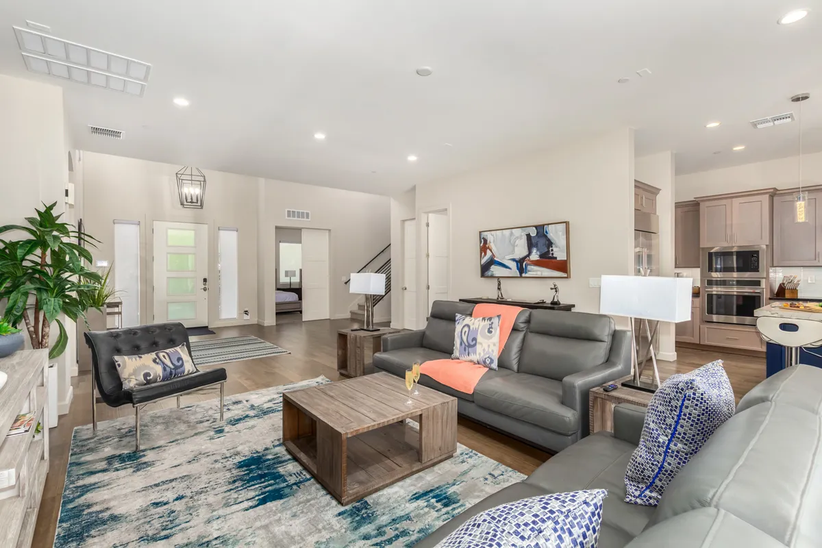 A bright and airy communal area with a sofa decorated with colorful cushions, a wooden coffee table, vibrant artwork, large windows, and decorative plants. The natural light and artful touches create a welcoming atmosphere for socializing or casual gatherings.